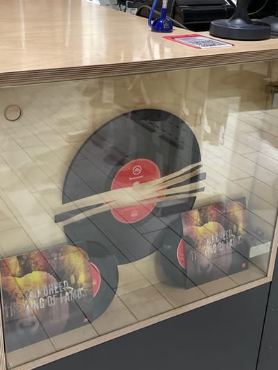 A glass-topped display case showcases a turntable and record sleeves. The turntable is a light-colored wood, with visible machinery and cables on the left side. Inside the case, several black vinyl records with red labels are arranged. Two album sleeves are also visible, featuring a design with yellow and brown hues, displaying the band name "LAMB OF GOD" and the title "WAKING OF LAMBS". Three thin, light-colored sticks rest on top of one of the records. The tabletop is wood panel.