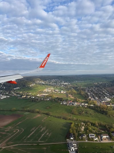 Aerial view from an EasyJet airplane over Berg, Luxembourg. The plane's wing is visible on the left. The landscape below features a mix of residential areas and green fields. The sky is partially cloudy.