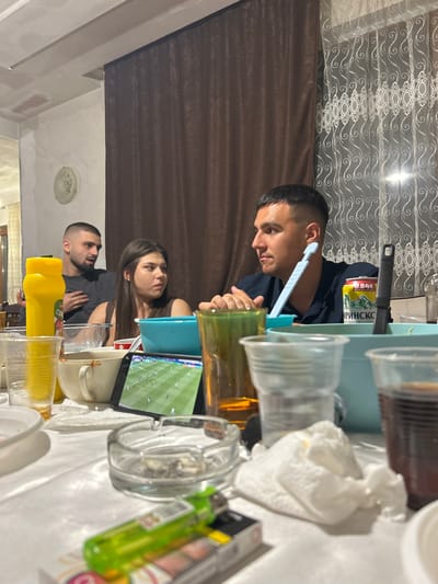 In Vaglen, Bulgaria, a group of people are gathered around a table, likely for a meal or social event. The scene depicts a casual indoor setting with a table covered by a white tablecloth. Several individuals are visible, including two men and a woman in the foreground and midground. One man, in the midground on the left, is wearing a dark t-shirt and has a short haircut. Beside him, a woman with long brown hair is looking to her right, wearing a dark sleeveless top. In the foreground on the right, a man with a short haircut and a dark blue shirt is looking to his left.

On the table, numerous objects are present. There are several clear plastic cups, some filled with dark liquid, and a bright yellow bottle, possibly a condiment. A blue plastic bowl and a white ceramic bowl are also visible. A smartphone is propped up, displaying what appears to be a soccer match. An ashtray with cigarette butts and a green lighter are in the foreground, suggesting smoking may be permitted in this area. Various other containers and plates are scattered across the table, indicating a meal has been or is being consumed. In the background, brown curtains are drawn, and a sheer white curtain with an ornate pattern hangs near a window. The overall impression is of a relaxed, informal gathering.