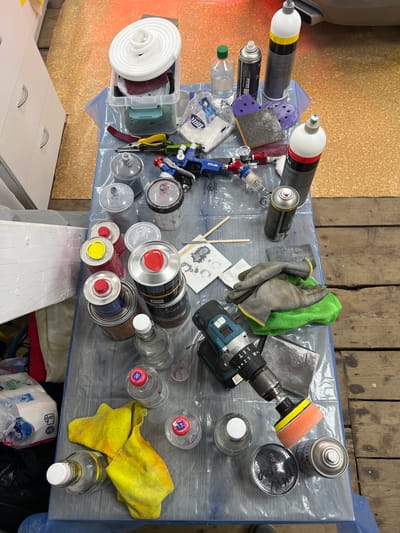 The image depicts a table covered with a blue plastic sheet, filled with various items used for automotive detailing or repair. These include spray cans, bottles of liquid, paint spray guns, sanding blocks, gloves, a power drill with a polishing attachment, and cloths. Several containers, some with red or yellow lids, are stacked on the table. In the background, a white cabinet with drawers and a portion of a car are visible. The table is placed on a wooden floor. The scene appears to be an indoor workspace, possibly a garage or workshop. The items suggest a task involving cleaning, polishing, or painting a vehicle.