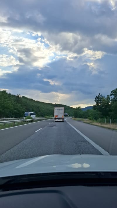The image depicts a view from a vehicle traveling on a highway. The highway is paved with asphalt and has white lane markings. On the left side of the highway, there is a guardrail and a densely vegetated hillside. On the right side, there is also vegetation and a fence. The sky is filled with dramatic, cloudy formations, with patches of blue sky visible between the clouds, suggesting either the early morning or late afternoon.
In the distance, a white truck with the logo "SONMEZ" is visible in the right lane of the highway. Further ahead, in the left lane, a white bus is present. The landscape surrounding the highway is hilly and covered with lush green trees. The perspective suggests the vehicle is moving forward. The foreground shows the hood of the vehicle from which the photograph is taken. The image does not show any specific landmarks that would allow for precise location identification within Gorno Varshilo, Bulgaria.
