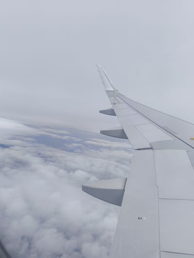 The image depicts an aerial view from an airplane window, showcasing a vast expanse of white clouds below with hints of blue sky peeking through. The right side of the frame is dominated by the wing of the airplane, a clean, white structure with visible panel lines and some markings, including the number "2" with two dots above it. The wing's upturned wingtip is prominently featured against the cloudy backdrop. The scene is devoid of any human presence or recognizable city landmarks, and therefore, cannot be associated with Bac Ai, Vietnam.