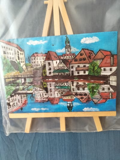 This is a painting of a city scene, likely České Budějovice, Czechia. The painting depicts a picturesque town with traditional European architecture, characterized by red-tiled roofs and white buildings with dark windows. A prominent clock tower with a green spire rises above the rooftops, suggesting a historical or central part of the city. The buildings are situated along a body of water, which reflects the architecture and the bright blue sky with scattered white clouds. A bridge spans the water in the mid-ground, connecting different parts of the town. Several boats are visible in the water, some appearing to be moored. The overall impression is one of a sunny day, with clear skies and a peaceful atmosphere. The painting is displayed on a small wooden easel, with the canvas wrapped in clear plastic.