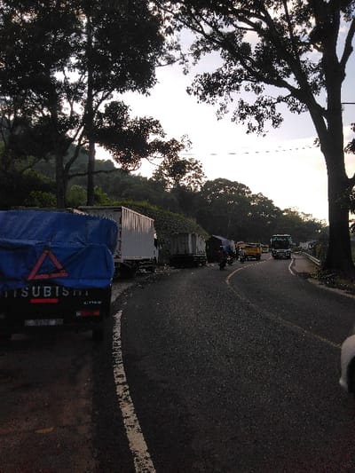 This image captures a daytime scene on an asphalt road in Cugenang, Indonesia, observed from a low angle. The road, which features a faded white solid line on the left shoulder and a partially visible dashed yellow center line, curves to the right in the mid-ground.
In the immediate foreground on the left shoulder, a dark-colored Mitsubishi pickup truck is stationary. Its bed is covered by a blue tarp, secured with ropes, and displays a prominent red triangular hazard symbol. Behind this truck, a white box truck and another dark-colored truck, also appearing stationary, are positioned on the left shoulder.
On the main road, several vehicles are present. At least two motorcycles are visible, one in the mid-ground approaching the curve from the right. Further down the road, a line of larger vehicles includes a yellow truck and at least two semi-trailer trucks (one with a green cabin, one with a white cabin), indicating traffic or a queue.
The environment is characterized by dense foliage. Large, mature trees with dark silhouettes dominate the left and right sides of the frame, their branches extending towards a light, possibly overcast, sky. Beyond the immediate roadside, the terrain appears hilly and is heavily covered in green vegetation. A white guardrail is visible on the right side of the road in the distance. No discernible human figures are clearly visible outside of vehicles.