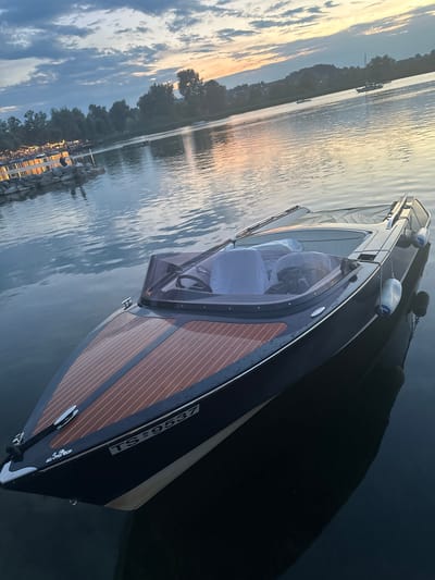 A dark-hulled motorboat with a wooden deck is moored on a body of water at dusk. The boat has a tinted windshield and the interior seats are covered with white fabric. The boat's registration number, "TS 9537", is visible on the hull. In the background, the shoreline is lined with trees and lights from a pier or waterfront establishment. Other boats are visible on the water, and the sky is filled with clouds illuminated by the setting sun. The scene is located in Rimsting, Germany, on the Chiemsee lake.