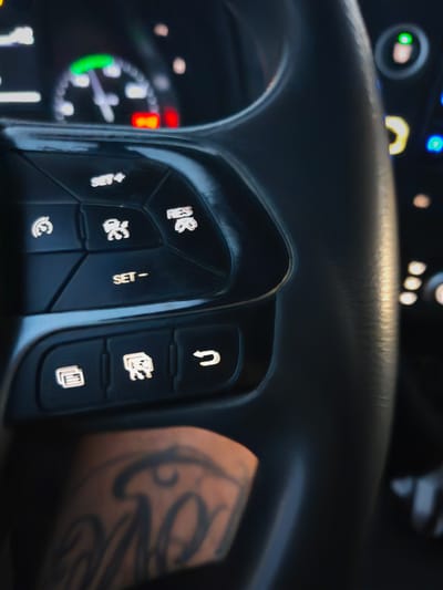 In this close-up, vertically oriented image, the focus is on the steering wheel controls of a vehicle, with a portion of the dashboard and a tattoo on a person's arm visible. The steering wheel is a dark, matte gray material. On the left side of the wheel, a cluster of illuminated buttons is present. The top button displays "SET+". Below it, there are two circular buttons with icons: the left one shows a stylized car with wavy lines indicating movement, and the right one depicts a car with a person inside. Further down, a button labeled "SET-" is visible. At the bottom of this cluster, two more illuminated buttons are seen. The left button has an icon resembling stacked papers or documents, and the middle button shows a car with two people inside. To the right of these, a button with a U-turn arrow icon is illuminated.
The dashboard in the background is blurred, but a speedometer with a green arc and a red indicator is discernible. The lighting suggests it is daytime, with light reflecting off the steering wheel. A tattoo, featuring elaborate script lettering, is visible on the person's forearm in the lower left corner of the frame, partially obscured by the steering wheel. The overall setting appears to be the interior of a car, potentially in Torrent, Spain, given the prompt. No specific actions or interactions are depicted, other than the presence of the controls.