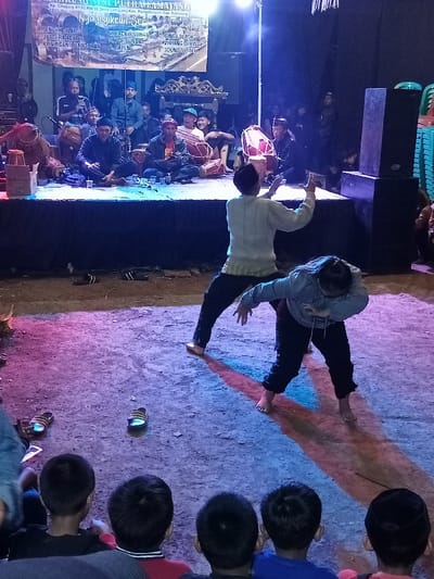 The image depicts a vibrant night-time cultural event taking place in Kp. Cikajang, Desa Lamajang, Pangalengan, Bandung, Indonesia. A raised stage is brightly lit, where several men are seated, playing traditional Indonesian drums (kendang) and other instruments, forming a musical ensemble. Above them, a banner with the title "PUTRACTAMAJANG Nyayogikeun Seni" lists various performances, including "PENCAK SILAT," "KACAPI SULING," "POP SUNDA," "SINGA DEPOH," and "WAYANG DAKWAH," suggesting a diverse program of traditional and contemporary arts.

In front of the stage, two barefoot individuals, a male and a female, are actively performing what appears to be a traditional dance or martial arts routine, likely Pencak Silat, illuminated by dynamic blue, red, and purple spotlights on the bare ground. Large speakers are visible beside the stage, indicating a sound system is in use. Several pairs of slippers are casually left on the ground near the performers. In the foreground, the backs of young boys are visible, forming part of an engaged audience watching the performance. The scene conveys a lively community gathering focused on cultural arts and music under the night sky.