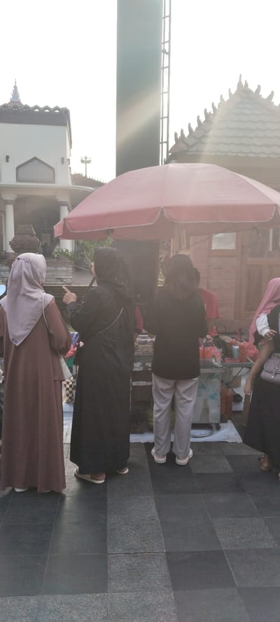 In the city of Kauman, Indonesia, a group of people stand near a street food stall under a large red umbrella. The sun is low in the sky, casting long shadows and creating a bright, hazy atmosphere. In the foreground, three women are visible from the waist down, two of whom are facing away from the camera, towards the stall. One woman, wearing a light brown hijab and a long brown dress, stands to the left. Next to her, a woman in a black hijab and black dress points towards something, possibly at the stall. To the right, a person in grey pants and a black top stands with their back to the camera. Behind them, a food cart with various bottles and containers is visible. In the background, traditional Indonesian architecture with distinctive tiled roofs can be seen, along with a tall, dark structure that resembles a tower or a part of a building. The ground is paved with dark grey tiles.