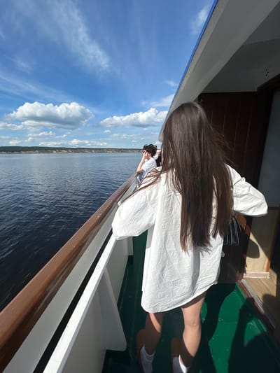 In Usol'e, Russia, on a bright, sunny day, a young woman with long, dark brown hair stands on the deck of a boat, facing away from the camera. She is wearing a loose-fitting white shirt and white socks with brown sandals. Her hair is blowing gently in the breeze. In the background, the dark blue water of a wide river stretches towards a lush green, tree-lined bank with sandy cliffs. The sky above is a vibrant blue, dotted with fluffy white clouds. Another person is visible in the background, leaning on the railing. The boat's deck is painted green, and there is a wooden railing with a white trim. Sunlight casts shadows across the deck.