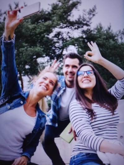 Three people are taking a selfie outdoors. The person on the left, a woman with blonde hair wearing a denim jacket over a white t-shirt, holds up a smartphone. To her right is a man with dark hair and a beard, wearing a blue and white checkered shirt over a white t-shirt. On the far right is a woman with long dark hair and glasses, wearing a striped long-sleeved shirt. She is making an "okay" gesture with her right hand. The man is also making an "okay" gesture with his right hand. They are all smiling. The background consists of trees and an overcast sky. The location is not identifiable as Balakliya, Ukraine.