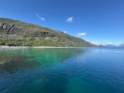 This media file is a landscape photograph taken in Lødingen, Norway, under a clear blue sky with a few wispy clouds. The scene depicts a body of water in the foreground, with a mountain range covered in greenery and rocky outcrops forming the backdrop. The water transitions from a shallow, turquoise hue near the shore to a deeper blue as it extends into the distance. Sunlight illuminates the water, revealing the rocky seabed in the shallower areas.
Along the shore, a road is visible, with several vehicles parked, including a white van and a dark-colored car. Trees and vegetation line the base of the mountain, creating a lush green band between the water and the rocky slopes. The mountain itself is characterized by steep, rugged terrain with scattered patches of vegetation.
In the far distance, other mountains can be seen, some capped with snow, suggesting a cool climate or higher altitude. The overall impression is one of natural beauty and tranquility, with a sense of open space and a bright, sunny day. The time of day appears to be mid-morning or early afternoon, given the strong sunlight and clear sky. There is no text visible in the image.