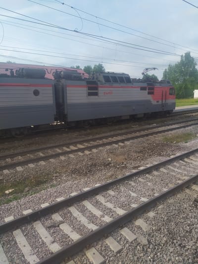 A side view of a Russian Railways electric locomotive, painted in gray and red, is shown on train tracks in Bol'shoi Dvor, Russia. The locomotive has a circular window on its side and is connected to overhead power lines via a pantograph. Trees and a red-roofed building are visible in the background. The foreground shows multiple sets of train tracks with gravel ballast and concrete sleepers. The sky is overcast with a few scattered clouds. There are no people visible in the image.