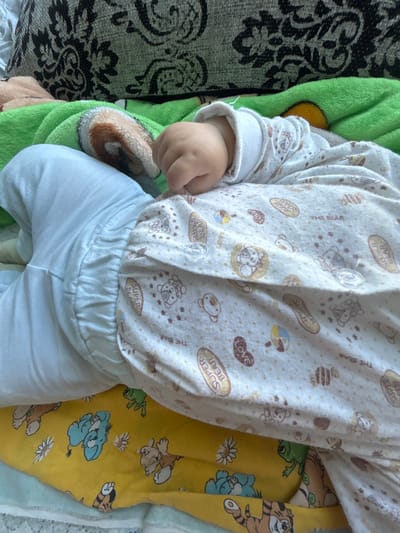 The image shows a close-up view of a baby lying on its stomach. The baby is wearing a white onesie with a pattern of cartoon bears, stars, and the words "SUPER BEAR" and "THE BEAR" on it. The baby's right arm is bent at the elbow and its fist is clenched. The baby is lying on a yellow blanket with cartoon images of elephants, frogs, and tigers. A green blanket is visible above the yellow blanket, and a patterned cushion is in the background. The scene appears to be indoors, on a bed or sofa. There is no indication of the location in Baymak, Russia.