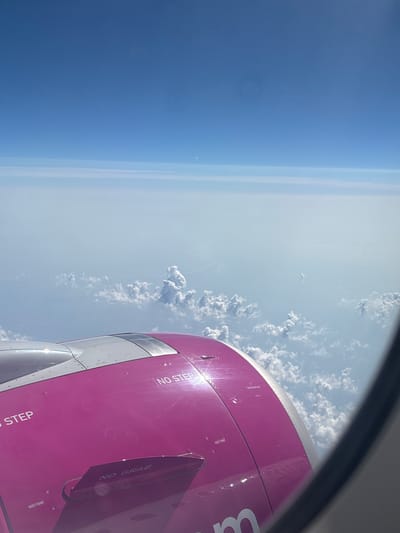 An aerial view from an airplane window shows a bright pink airplane engine against a backdrop of clouds and a clear blue sky. The engine housing is prominent in the foreground, with the words "NO STEP" and "NO GRAB" visible. The clouds are scattered below, with a hazy layer of mist or thinner clouds in the distance, obscuring the view of the ground. The sky is a vibrant blue, transitioning to a lighter shade near the horizon. There is a faint sliver of the moon visible in the upper part of the sky. The image provides no information about the city of Ub Municipality, Serbia, or any people.