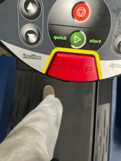 The image depicts a close-up, high-angle view of a person's lower left leg and foot on a treadmill, with a significant portion of the treadmill's control panel visible in the upper half. The individual is wearing light-colored, possibly white or light grey, loose-fitting pants and a light brown or tan shoe on their left foot, which is positioned on the black running belt of the treadmill. The control panel is grey and black, featuring several buttons and labels. Prominently displayed is a large red "STOP" button with a yellow border. To its right, a green triangular "start" button is labeled "quick" and "start." Above these, a red circular button with a white geometric symbol is present. On the left, two grey circular buttons with up and down arrows are positioned above the label "incline." The term "speed" is partially visible on the right side of the panel. A blue floor mat or covering is discernible at the top left and bottom right corners of the frame. The scene is located within the city of Covilhã, Portugal.