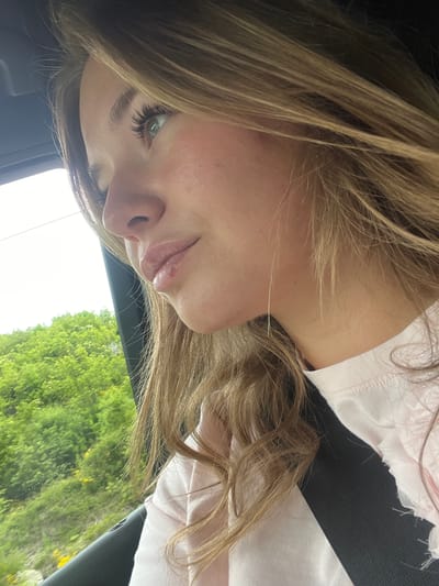 A young woman is photographed in a car in daylight. She is wearing a light pink shirt and a black seatbelt. She has blonde hair and visible eyelashes. Outside the car window, there is bright green vegetation. The location is not discernible based on the provided visual details.