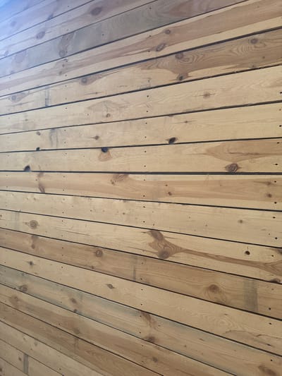 The image displays a close-up, angled view of a wall constructed from horizontal wooden planks. The planks are light brown in color with visible wood grain, knots, and some minor imperfections such as small holes and darker streaks. The texture is rough, suggesting unfinished or rustic wood. The planks are uniformly spaced, creating distinct horizontal lines across the frame. The lighting suggests an indoor environment with ambient light illuminating the textured surface. The image does not depict any people, identifiable landmarks, or contextual environmental details that would allow for the determination of its location in Otradny, Russia.
