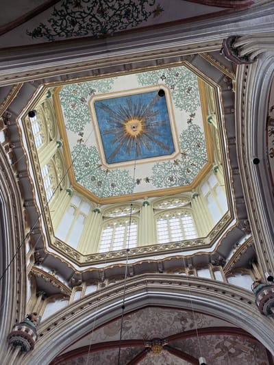 The image shows a low-angle view of the ornate ceiling and upper walls of a gothic cathedral in 's-Hertogenbosch, Netherlands, identified as the Sint-Janskathedraal (St. John's Cathedral). The architecture features ribbed vaults, pointed arches, and intricate decorative elements.
The central focus of the ceiling is an octagonal section painted a vibrant blue, with a prominent symbol of the Eye of Providence (an eye within a triangle, surrounded by rays of light) at its center. This symbolic eye is likely a representation of God. Surrounding the blue octagon is a border of white adorned with green, floral, and vine-like patterns. The date "1840" is visible within the decorative border in the upper portion of the image.
The upper walls are punctuated by tall, arched windows with Gothic tracery, letting in diffused light. The exterior of the octagonal section of the ceiling is supported by cream-colored columns and architectural features, with decorative corbels or sculpted heads visible in some sections. Dark, thin wires, possibly for lighting or sound systems, hang down from the ceiling. The overall impression is one of grandeur and historical significance, characteristic of major European cathedrals.