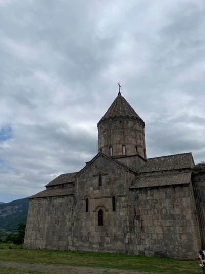 The image displays the Tatev Monastery, situated in Tatev, Syunik Province, Armenia. The scene features the monastery's main church, constructed from light to dark gray cut stone blocks. A prominent circular drum, topped by a conical roof with a small dark cross at its apex, rises from the central structure. The drum features several narrow, vertical rectangular windows. Below the drum, gabled roof sections extend, supported by thick stone walls with narrow, arched windows. The roof tiles appear to be made of dark, possibly weathered, stone.

In the foreground, a patch of uneven green grass occupies the bottom portion of the frame. The background on the left reveals distant, undulating mountains or hills with some green foliage. The sky is largely overcast, dominated by varying shades of grey clouds, with a small patch of lighter sky visible on the far left.

At the bottom right corner of the frame, two individuals are partially visible. One person wears a white top and dark pants, facing away from the camera, while the other is mostly obscured. They are standing near the base of the monastery wall. No specific actions or interactions are discernible.