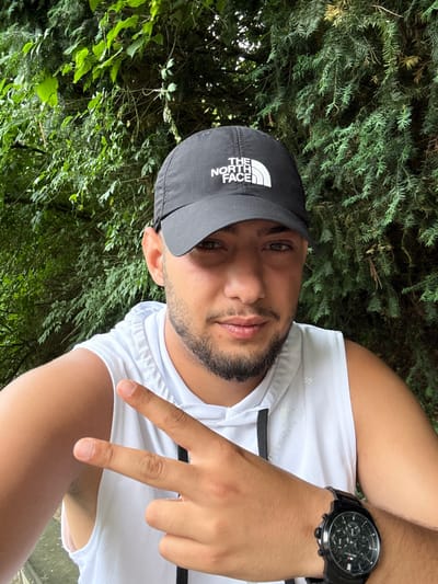 A man is pictured from the chest up, wearing a black "The North Face" baseball cap, a white sleeveless athletic shirt, and a black watch. He is giving a "peace sign" with his right hand. He has a short, dark beard and appears to be of Middle Eastern descent. The background consists of dense green foliage suggesting a park or wooded area. The location cannot be determined as Liestal without additional identifying details.
