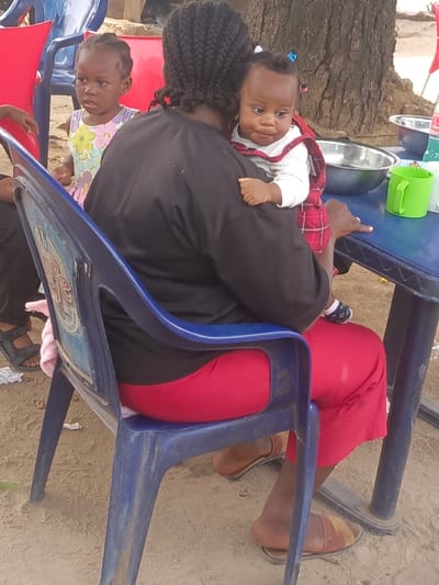 A woman with braided hair sits in a blue plastic chair wearing a black shirt and red pants. She holds a baby in a red and white plaid dress. Another young child in a floral dress stands beside the woman's chair. A blue table with a silver bowl and a green cup is beside the woman. The scene appears to be outdoors, possibly at a gathering. The ground is sandy.