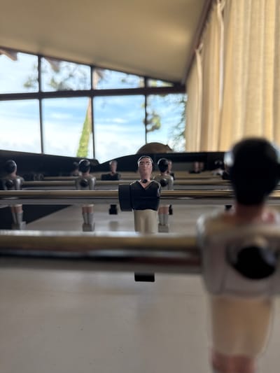 The image presents a close-up, low-angle perspective of a foosball table, initially rotated 90 degrees clockwise. When viewed correctly, a row of foosball player figures on their metal rods extends horizontally across the frame. The figure in the immediate foreground, dressed in a black top and white bottom, is sharply in focus, facing towards the left. Subsequent player figures and rods further down the table are progressively blurred.
The background reveals a large window with dark frames, offering a glimpse of the outdoors. Through the window, a bright blue sky with scattered white clouds is visible, along with blurred green foliage, indicative of a pleasant natural landscape. This natural light streaming in suggests the time of day is daytime.
Above the foosball table, a light-colored ceiling with horizontal blinds or slats is visible, also out of focus. A plain, light-colored wall forms the right side of the indoor setting. No specific activity or game play is depicted, only the static setup of the foosball table. There is no visible text in the image. The scene conveys a relaxed, recreational atmosphere within an indoor setting, likely in Lège-Cap-Ferret, France.