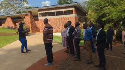 A group of approximately ten people stand outdoors on a brick walkway near a brick building. One man in a dark blue shirt and blue pants appears to be speaking and gesturing towards the building. The others, mostly men, are facing him, dressed in a mix of casual and formal attire. Trees are visible in the background, suggesting a park-like or campus setting. The overall impression is a tour or presentation of the building.