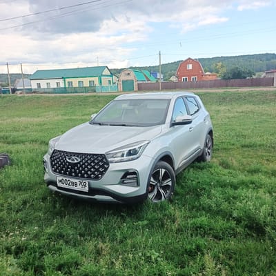 In Podlubovo, Russia, a silver Chery Tiggo 4 Pro SUV is parked on a grassy field with a village in the background. The SUV, with license plate M002BB 702, faces slightly to the left, showcasing its front grille and headlights. Behind the vehicle, a row of houses with green roofs and fences lines the edge of the field, leading up to a forested hill. The sky is overcast with scattered clouds. The environment appears rural and residential.