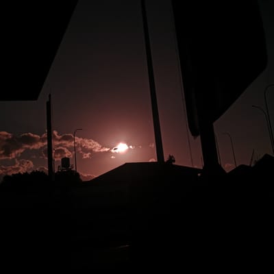 This image captures a sunset over Sihomlhok, Indonesia, with a dramatic interplay of light and shadow. The sky is a deep, rich reddish-orange, indicative of a setting sun. The sun itself is partially obscured by clouds, creating a bright, luminous halo effect. Wispy, pink-tinged clouds are scattered across the sky.
In the foreground, silhouetted against the vibrant sky, are various man-made structures. To the left, a large, dark, angular object dominates the upper portion of the frame, possibly part of an overpass or building. Further to the right, another tall, slender structure, likely a utility pole or communication tower, stands prominently.
Below these structures, a dark silhouette of a rooftop is visible, possibly belonging to a house or other building. Near the center-left, a water tank on a metal frame is perched atop a silhouetted tree line, suggesting residential or industrial infrastructure. On the right side of the image, several streetlights, also in silhouette, are positioned along what appears to be a road.
The overall scene is characterized by deep shadows and silhouettes, emphasizing the radiant colors of the sunset. The image conveys a sense of urban landscape at dusk, with the natural beauty of the sky contrasting with the stark outlines of human development.