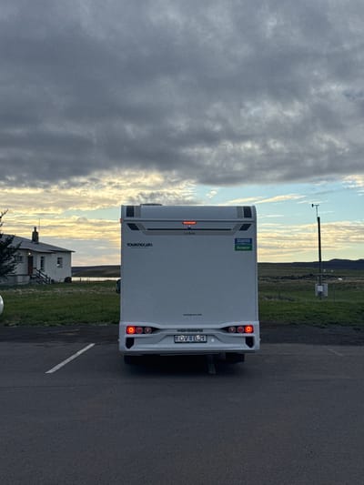 The image, taken in Reykjahlíð, Iceland, shows the rear of a white camper van parked on an asphalt surface. The camper van is facing away from the viewer, with its red taillights illuminated. Visible text on the back includes "TOURINGCARS" with "MOTORHOME RENTAL" below it, and "WEINSBERG" with "touringcars.com". The license plate reads "QV B21". A sticker displays "BÍLALEIGA Akureyrar" (Car Rental Akureyri) and the "Europcar" logo. Below a horizontal light strip, the word "CaraSuite" is visible.

The scene depicts a rural or semi-rural landscape under a dramatic sky. Green fields stretch into the distance, with what appears to be a body of water or lake visible further back, flanked by distant hills or mountains. To the left, a white house with a dark roof and chimney is situated in the mid-ground. On the right, a tall wooden pole with attached equipment, possibly a camera or sensor, stands against the horizon.

The time of day is either early morning or late evening, characterized by soft, warm, golden light illuminating parts of the sky and clouds. The sky is partly cloudy, with dark, grey clouds dominating the left side, contrasting with brighter, golden-streaked clouds towards the right, suggesting a sunrise or sunset. The overall atmosphere is peaceful and tranquil.