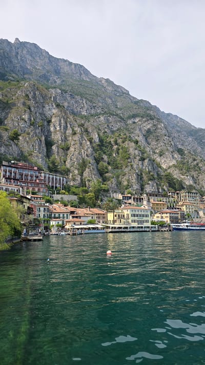 The image depicts a waterfront scene in Limone sul Garda, Italy, with a steep, rocky mountain rising sharply behind a cluster of buildings. The buildings, varying in color and architectural style, are situated along the shoreline, some appearing to be hotels or restaurants with outdoor seating areas. A body of water, likely Lake Garda, occupies the foreground, its surface reflecting the surrounding landscape with a greenish hue and visible ripples. Several boats are docked or in the water near the shore, including a larger passenger ferry. A few individuals are visible on piers and along the waterfront. The overall atmosphere is one of a scenic, European lakeside town.