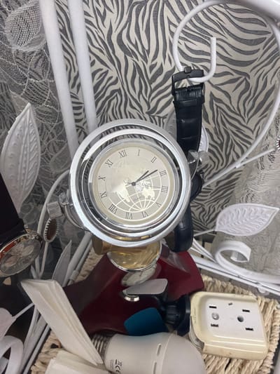 The image displays a close-up view of a collection of items, seemingly in a state of disarray, within a room. The backdrop features a distinctive black and white zebra-print wallpaper and a sheer white lace curtain. Prominently featured is a silver-toned analog clock with Roman numerals, a black leather watch strap, and a portion of a black leather watch band. Several decorative white leaf-shaped elements and white cylindrical rods are also visible, possibly part of a larger decorative structure or piece of furniture.

Other objects in the scene include a white light bulb, a cream-colored electrical outlet cover with two sockets, and a woven basket. The overall impression is one of accumulated belongings and possibly a crafting or storage area. The image does not provide any contextual information to suggest a specific location in Khyrdalan, Azerbaijan.