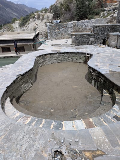 The image depicts an outdoor scene in the city of Panamik, India, focusing on a partially constructed or renovated outdoor pool area. The pool itself is empty and filled with a muddy, sandy substance, with its stone-lined walls visible. The surrounding area is paved with irregular, flat stone tiles, some of which appear to be in the process of being laid or repaired, with exposed mortar and debris.
In the background, a traditional building with a flat roof and windows is visible, and a person dressed in dark clothing with an orange tool in hand is walking along the edge of the pool area. Further in the background, a rocky, mountainous landscape under a cloudy sky suggests a rural or mountainous setting. Stone walls and structures, some appearing aged and others more recently built with smaller stones, are integrated into the hillside, characteristic of the region's architecture. The overall impression is one of ongoing construction or maintenance in a remote, natural environment.