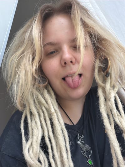 This media file is a close-up, rotated image of a person with blonde dreadlocks. The subject is looking towards the camera and sticking out their tongue, with one eye partially closed. They are wearing a dark shirt with a silver pendant that resembles a wolf or dog with a green gem. The background is out of focus and appears to be indoors with a light-colored wall or sheet. The lighting suggests it is daytime. The image is cropped to focus on the subject's face and upper torso, with their dreadlocks framing their head and shoulders. The overall impression is playful and informal. No text is visible in the image.