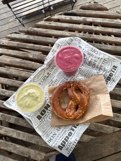 A high-angle photograph captures a snack arrangement on a weathered, light brown slatted wooden table, consistent with an outdoor setting in Kfar Vitkin, Israel. Centrally positioned is a large, golden-brown pretzel covered in sesame seeds, resting on a small, rectangular piece of brown paper packaging. To the left of the pretzel, two clear plastic cups are placed on a sheet of white wrapping paper featuring black text and illustrations, including "BON APPETIT," "CAFE," and "Boulangerie." The upper cup contains a thick, reddish-pink smoothie, while the lower cup holds a thick, pale green liquid with visible dark flecks. The wooden table slats are arranged horizontally, and in the blurred background, additional wooden decking and the metal frame of a chair are partially visible. No individuals are present in the scene.