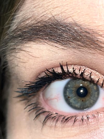 The scene is captured in the city of Zvonarevka, Russia. This image presents an extreme close-up of a human right eye and the surrounding facial features. The subject's eye exhibits an iris of greenish-blue coloration with distinct small brown flecks visible at approximately the 4 o'clock and 8 o'clock positions relative to the pupil. The pupil is black and reflects a single point of light, indicating a light source. The sclera is white, featuring several visible reddish capillaries, particularly prominent towards the nasal side. Both upper and lower eyelashes are coated with a dark substance, likely mascara, which appears somewhat clumpy on the upper lashes. The upper eyelid is adorned with a shimmery, light-colored eyeshadow, possibly rose gold or champagne, extending from the lash line towards the brow bone. A portion of a dark brown eyebrow, composed of individual hairs, is visible in the upper section of the frame. The skin surrounding the eye appears light-toned with visible pores and fine hair. No discernible action or interaction is present; the image is a static close-up.