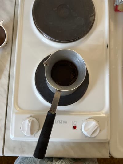 The image shows a stovetop in Nizhnee Sancheleevo, Russia, with a pot of coffee brewing. The stovetop is white and has two circular heating elements. One element is in use, with a metal pot containing dark liquid on it. The pot has a black handle. The stovetop has control knobs, one of which is visible with settings numbered 1 through 6. A small red light is illuminated on the control panel, indicating that the stove is on. The overall scene suggests a domestic setting.