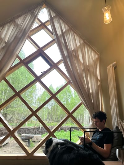 A man sits on a couch next to a dog, writing on a tablet. The man wears a black shirt and dark pants. The dog, primarily black with white markings, is partially visible, with its head near the window. The environment includes a room with a diamond-shaped lattice window through which a green, wooded area is visible. Beige curtains partially cover the window. A light fixture hangs from the ceiling, and a radiator is mounted on the wall. The location is not specified in the image.