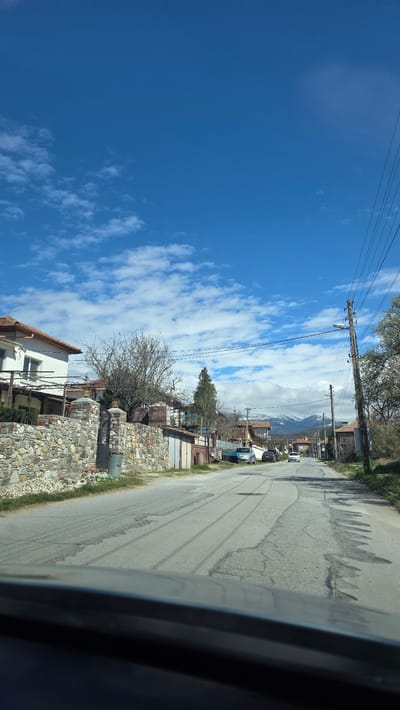 The image shows a street in the village of Slatino, Bulgaria. The view is taken from inside a vehicle, looking out the passenger window. A section of the interior trim of the vehicle is visible on the right. The street is asphalt, with visible cracks and patches. On the right side of the road are houses and fences made of stone and brick. Utility poles run along the right side of the street with wires overhead. In the distance, mountains are visible under a sky with scattered clouds. The sky is predominantly blue. The vegetation appears to be sparse, suggesting it may be early spring or late autumn. The houses are of varying sizes and styles, typical of rural construction. Several vehicles are parked along the street in the distance.