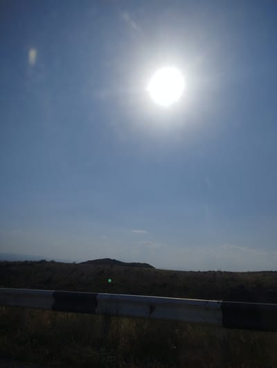 This is a landscape image taken in Dashtadem, Armenia. The scene is dominated by a bright, overhead sun, casting a strong glare and making it difficult to discern fine details in the sky, which is a clear blue with some wispy clouds. In the middle ground, there is a rolling, grassy landscape with a few small, dark hills. A section of a guardrail, painted with alternating black and white stripes, runs across the foreground, obscuring the lower part of the landscape. The overall impression is a bright, sunny day in a rural, open area.