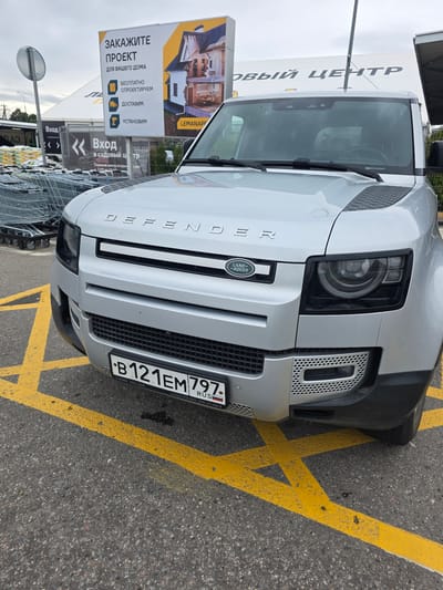 The image shows a silver Land Rover Defender parked on asphalt marked with yellow lines. The vehicle's license plate reads "B121EM797 RUS". Behind the vehicle, a large white banner with Russian text is visible, advertising a service called "LEMANAPE" related to "PROJECTS FOR YOUR HOME" including free design, delivery, and installation. To the left, shopping carts are stacked, and another banner reads "ВХОД В САДОВЫЙ ЦЕНТР" (Entrance to the Garden Center). The scene appears to be an outdoor commercial area. The specific location is identified as Sholokhovo, Russia. There are no people visible in the image.