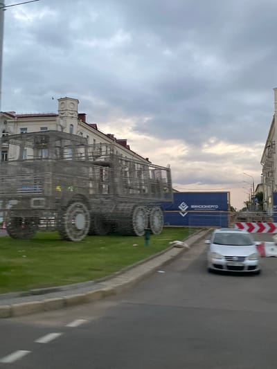 This media file shows an outdoor scene that appears to be in a city or town. The primary subjects are a large, wireframe sculpture resembling a bus or tram, a white car, and a building. The sculpture is elaborate and detailed, with what look like large wheels. It is positioned on a grassy area next to a sidewalk and a road. The white car is on the road, moving away from the camera. Behind the sculpture and to the right, there is a blue construction barrier with white text. The building in the background is multi-story with a distinct architectural style, including a tower-like structure on one side. The scene is set under a cloudy sky, suggesting it might be late afternoon or early evening. The overall impression is one of urban infrastructure with a public art installation.
The blue barrier has white text that reads "ВЯЛІКІЯ МЯДВЕДЗІ" which translates to "BIG BEARS" in Belarusian. This is a strong location cue, potentially indicating the name of the installation or a related event.
The angle of the shot is from a slightly elevated perspective, looking down and across the scene. The overall mood is somewhat subdued due to the overcast weather. The presence of a construction barrier suggests ongoing development or repair work in the area.