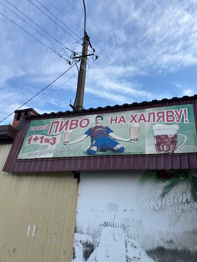 An advertisement for beer "on the house" in Ulyanovka, Russia, features an image of a soccer player holding a beer glass, likely soccer player Lionel Messi, on a sign attached to the roof of a building. The sign states "1+1=3," implying a special offer. An electrical pole is visible in the background against a partly cloudy sky. The building appears to be a simple, single-story structure with corrugated metal siding.