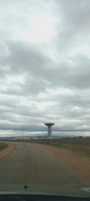 The image presents an exterior view captured from within a vehicle, depicting a rural or semi-urban landscape under an overcast sky in Sidi Slimane Moul Al Kifane, Morocco. The upper two-thirds of the frame are dominated by a dense, grey cloud cover, exhibiting brighter illumination near the top-center. The lower foreground shows the dark dashboard and windshield of a vehicle, with a small, circular dark object visible on the dashboard.
A two-lane paved road, appearing worn with visible cracks and patches, extends from the bottom of the frame, curving slightly to the left as it recedes towards the horizon. The road is bordered by uncultivated, dry, reddish-brown earth with sparse green and brown vegetation. In the mid-ground, a prominent, modern concrete water tower stands, characterized by a tall, cylindrical shaft supporting a wide, flat, disk-shaped reservoir structure. To the left of the water tower, a lattice-structured utility tower is partially visible in the distance. Beyond these structures, a cluster of low-rise, light-colored buildings is discernible against the horizon, which features faint, distant hills or terrain. No individuals, vehicles other than the perspective vehicle, or explicit actions are present within the frame.