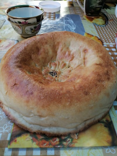 A large, round, flatbread, possibly a tandoori roti, is centered on a patterned tablecloth. The bread is golden brown and has a textured, slightly dimpled surface. A small cluster of black seeds, likely sesame seeds, is visible in the center of the bread. To the left of the bread, two decorative ceramic bowls are stacked, with floral patterns visible on the lower bowl. In the background, a white plastic lid and a portion of a dark mug with a picture on it are also visible. The tablecloth has a checkered and patterned design, with images of fruits and flowers. The setting appears to be outdoors, likely in a residential area in Gumbaz, Uzbekistan, given the traditional appearance of the bread and bowls.