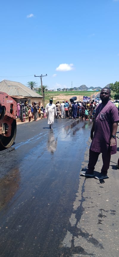 A group of people are gathered on a wet asphalt road, possibly in Kayarda, Nigeria, where roadwork is underway. A large steamroller is visible on the left side of the image, its red paint chipped in places and bearing the inscription "CC424HF". The wet road surface reflects the bright sky and the surrounding environment.
In the foreground on the right, a man in a deep purple traditional outfit with intricate embroidery on the chest is standing and looking directly at the camera. He is wearing grey athletic shoes. To his left, a man in a white flowing robe and a green cap is walking away from the camera, looking towards the crowd.
The crowd consists of individuals of various ages, many of them children, dressed in a mix of traditional and casual clothing. Some are observing the roadwork, while others appear to be interacting with each other. Several people are carrying items, including what appears to be a large black speaker. In the background, there are buildings, palm trees, and a utility pole. Further in the distance, rolling hills and a communication tower are visible under a clear blue sky with scattered clouds. The overall scene suggests a community event or a disruption caused by the road construction.