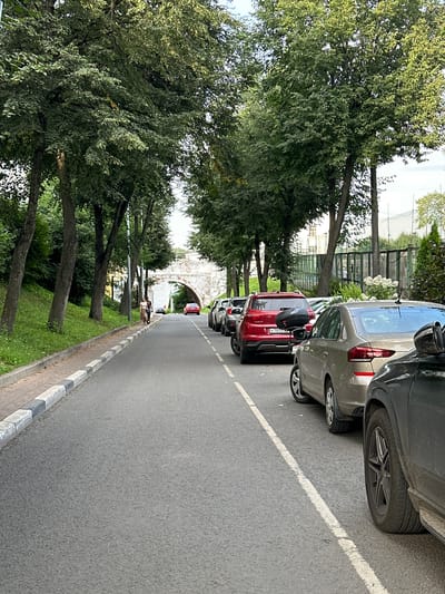 The image shows a street scene in Putyatino (Semivragovskaya S/A), Russia. Tall trees line both sides of the asphalt road, creating a shaded canopy. On the left side of the road, there is a grassy slope with more trees. On the right, several cars are parked along the street, and a line of more cars is visible further down the road. In the distance, an arched stone bridge is prominent. Two people are walking on the sidewalk on the left side of the road, near the bridge. The overall impression is a quiet street with a natural, green environment.