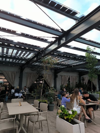 The image depicts an outdoor or semi-enclosed dining area within a restaurant or cafe located in Oława, Poland. The space features a modern industrial design with a substantial overhead structure composed of dark grey steel I-beams, supporting a grid of transparent panels that integrate photovoltaic cells or light-filtering elements, allowing natural light. Steel tension cables provide additional structural support.
The walls in the background are light grey, textured, and several large openings are covered with draped, beige fabric curtains. The flooring consists of light brown, horizontally laid planks, resembling a wooden deck. Various potted plants and small trees are distributed throughout the area, including a prominent white rectangular planter in the foreground containing a flowering plant with white and pink blooms.
Numerous light-colored, modern dining tables, both rectangular and round, are arranged across the space. These tables are accompanied by matching light beige/brown molded chairs with perforated backs. Approximately 15-20 individuals are visible, seated at these tables.
In the foreground to the right, a young male with dark hair and arm tattoos is seated, wearing a black t-shirt and shorts, actively eating. Opposite him, a young female with long brown hair, wearing a striped white top and white shorts, is also seated and eating. A young child in a blue long-sleeved shirt is seated to the female's left at the same table. Further back, on the left, a group of approximately five adults is seated at a table, engaged in conversation. Other patrons are visible in the mid-ground and background, seated at tables, eating, or socializing. An unoccupied rectangular table with a small, light-colored menu holder or table number stands in the immediate foreground.