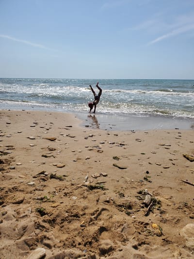 A person performs a handstand near the waterline on a sandy beach. The person is wearing dark patterned shorts and a red cap. The sea is calm with small waves lapping at the shore, and the sky is clear and blue. The beach is covered in sand, small pebbles, shells, and some seaweed. The location is the coast of Chornomorske, Ukraine.