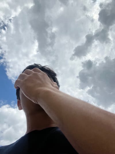 A low-angle, medium shot captures a person's head and arm from behind, looking up towards a cloudy sky. The person is wearing a black t-shirt and has dark hair. Their right arm is raised, with their hand covering their face and forehead. The background consists of a mix of blue sky and large, gray, cumulus clouds. The lighting suggests an overcast day. The image is taken outdoors, with no identifiable urban or natural landmarks, making it impossible to ascertain the location.